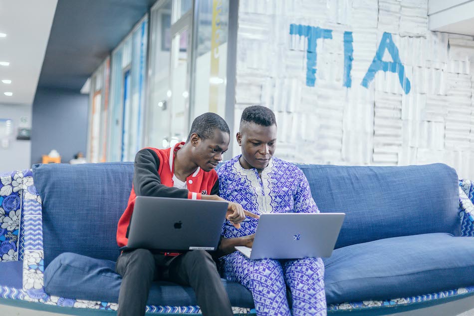 Two male engineers sitting together on a blue sofa, working on laptop computers (Andela, Annual Letter, CZI).