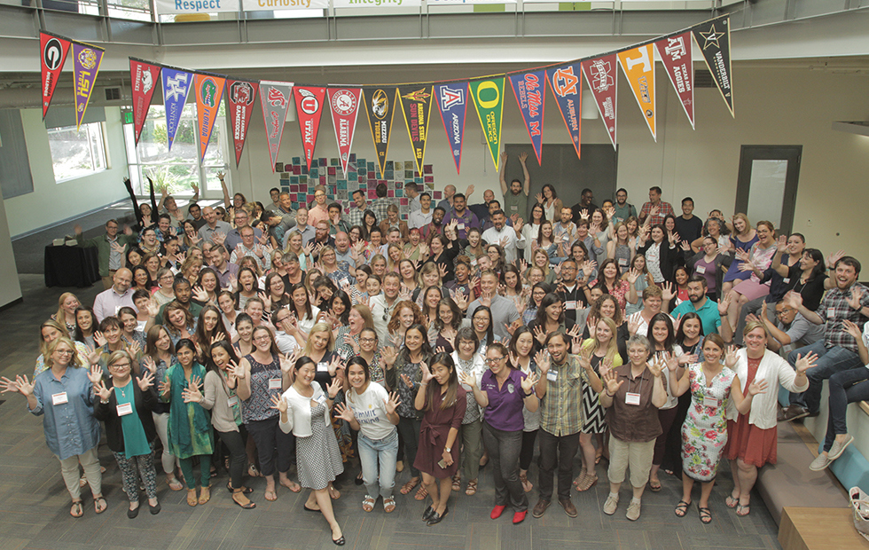 Large group of smiling teachers showing open hands at a conference, beneath a hanging line of college pennants (Summit Learning, Annual Letter, CZI).