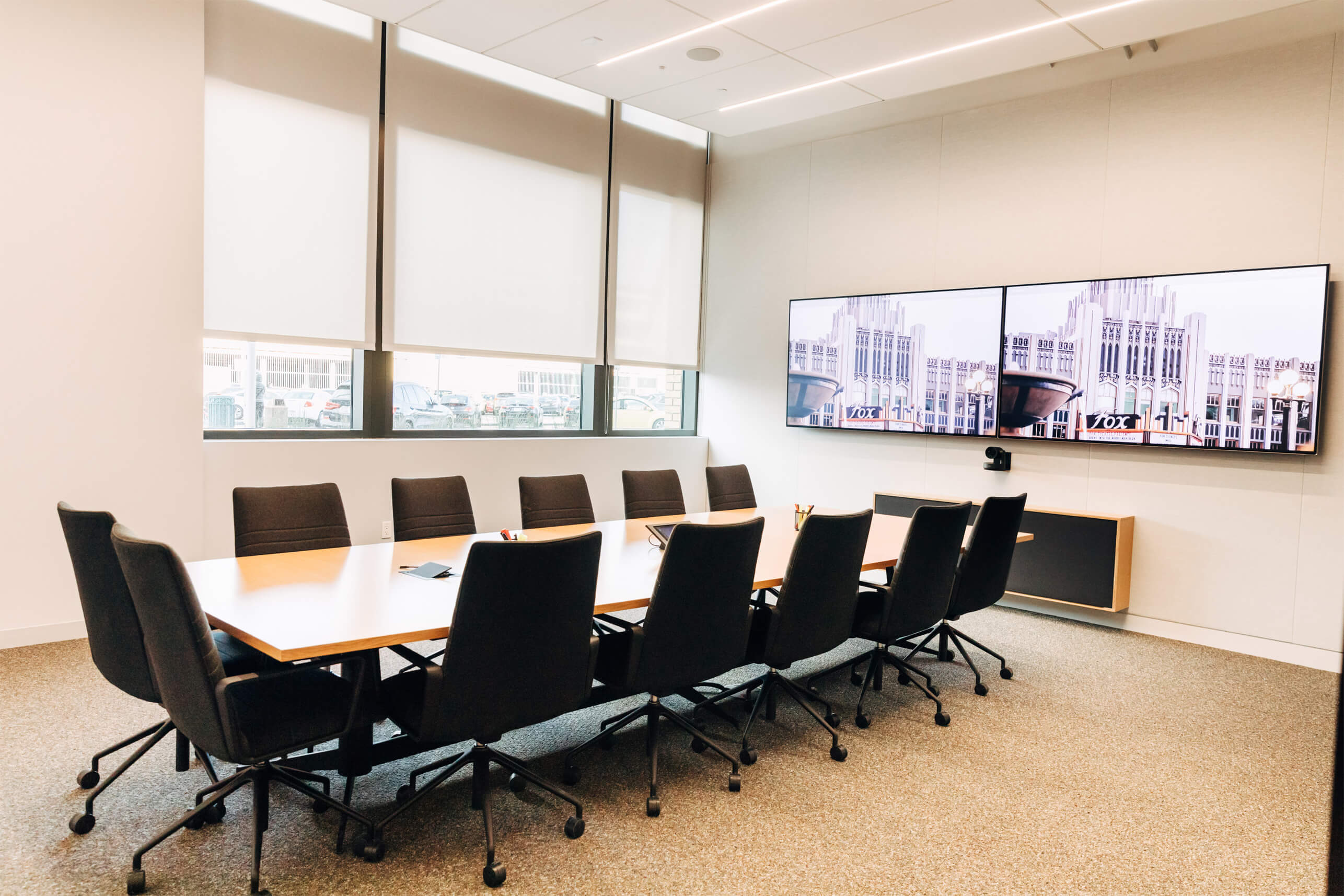 Conference room with a table and twelve chairs, two digital screens and windows.