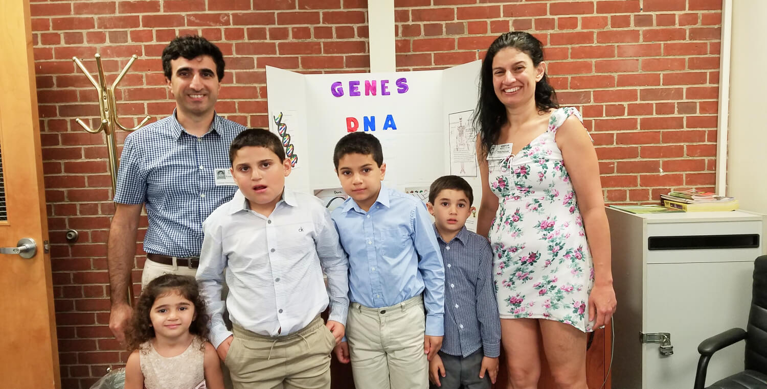 Emile Najm (far left), four children (middle), and Emile’s wife (far right) pose in front of a poster about genes and DNA