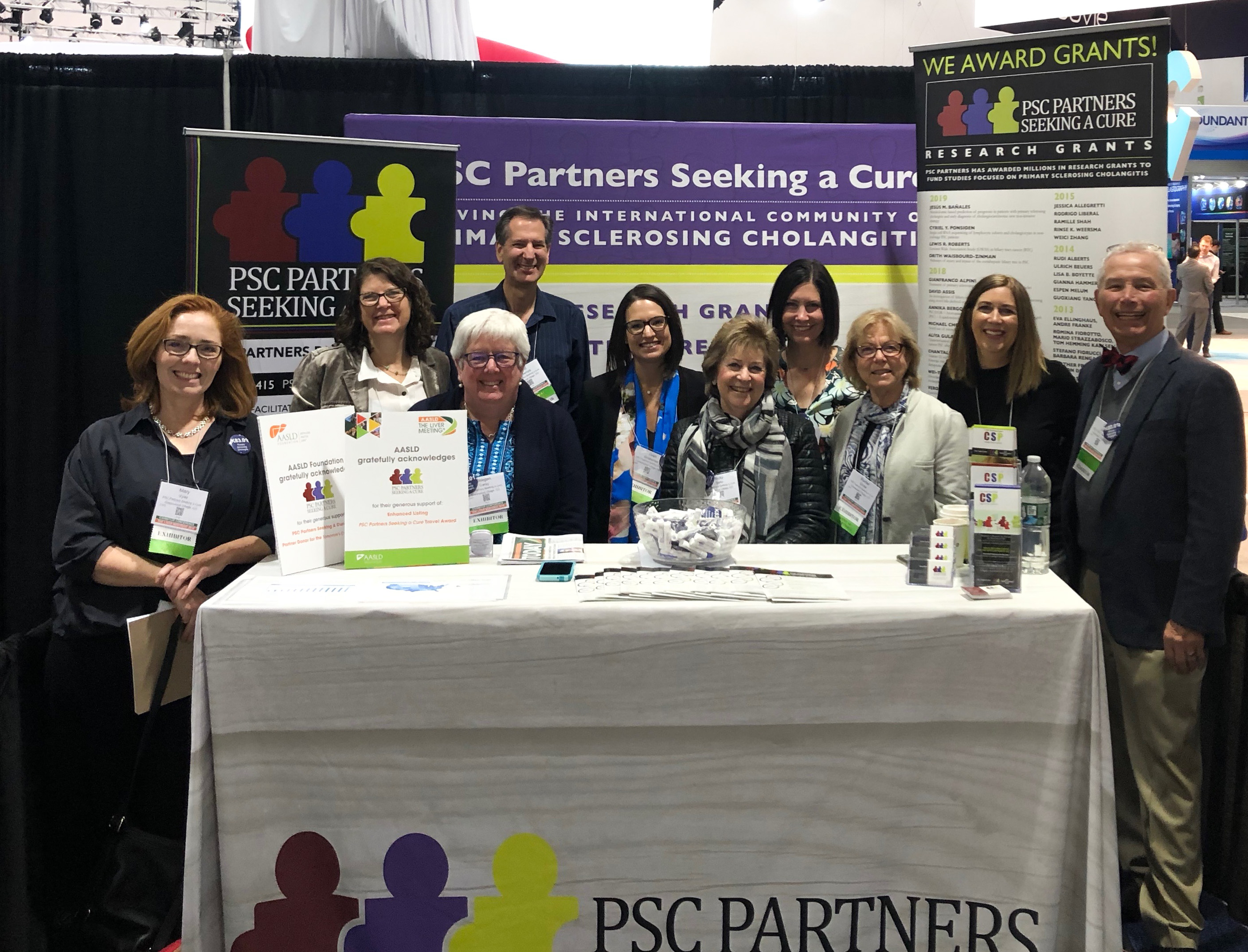 A group of 10 men and woman stand at a booth at the 2019 Liver Meeting in Boston