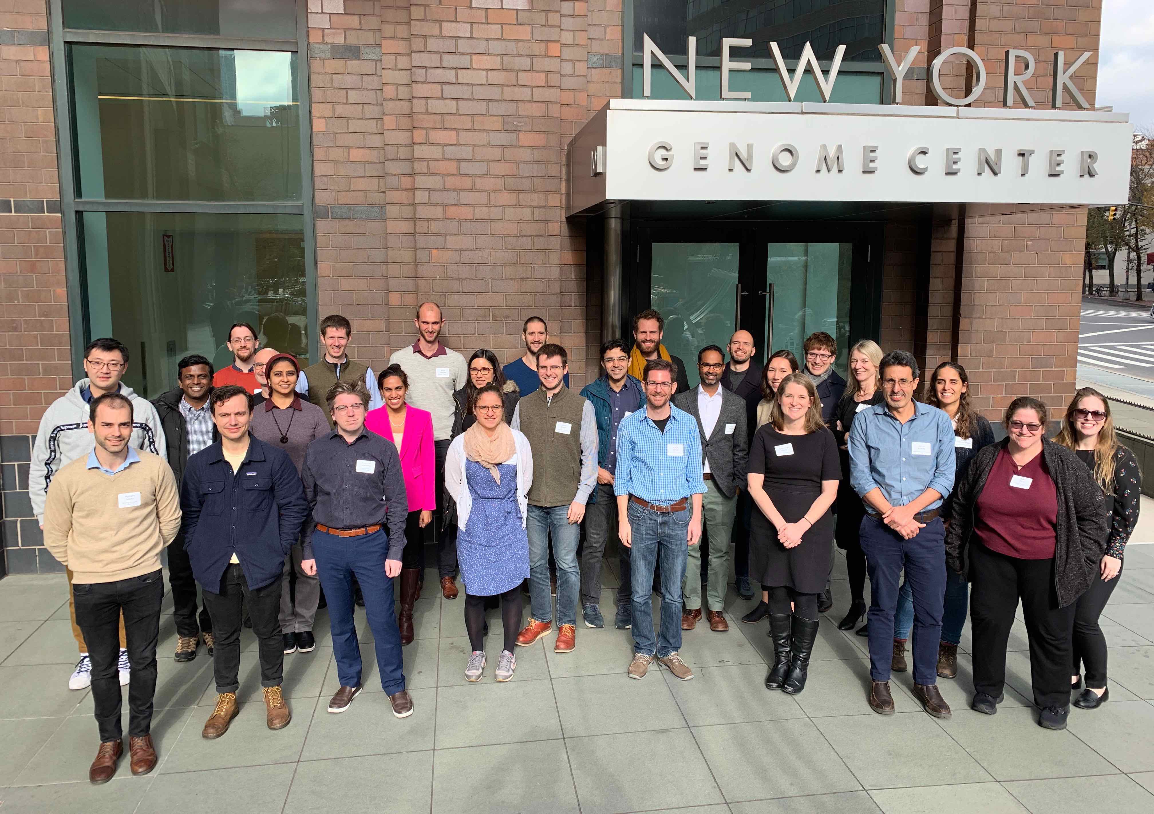 A group of attendees at the normjam workshop pose for a photo outside a building sign that reads New York Genome Center.