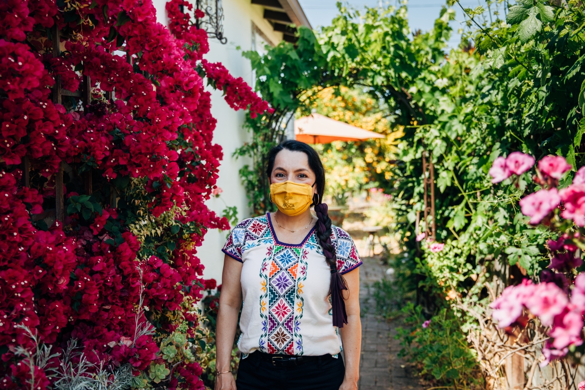 A woman in a colorful blouse, wears a yellow face mask bearing the NFOCA logo in her garden.