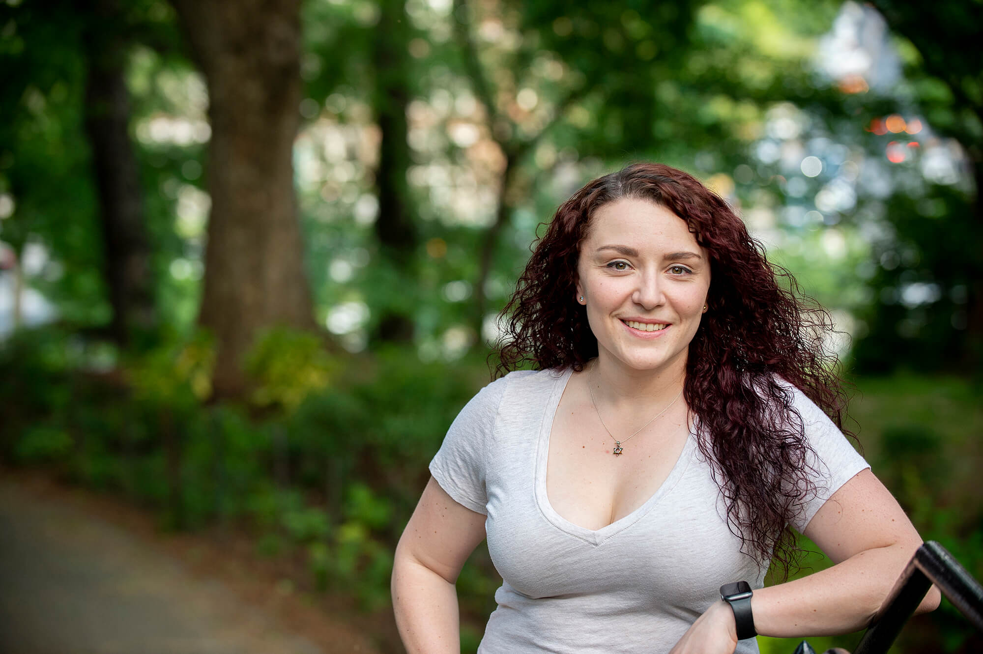 A woman wears a white t-shirt and smiles. Trees are blurred in the background.