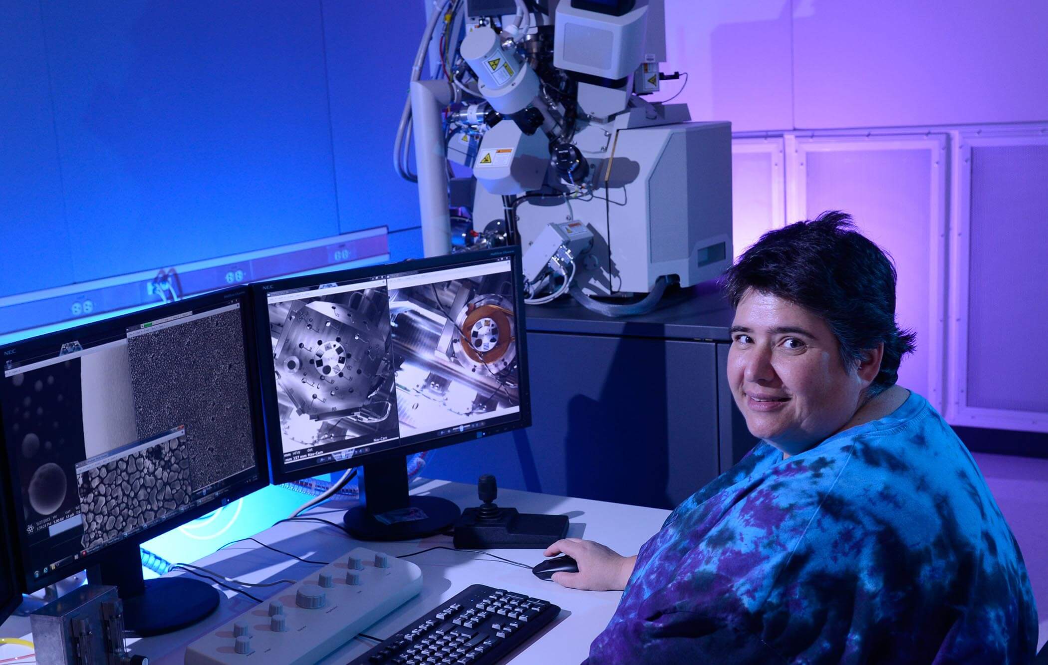 A women looks at the camera and smiles as she work at two monitors in a blue-lit room.