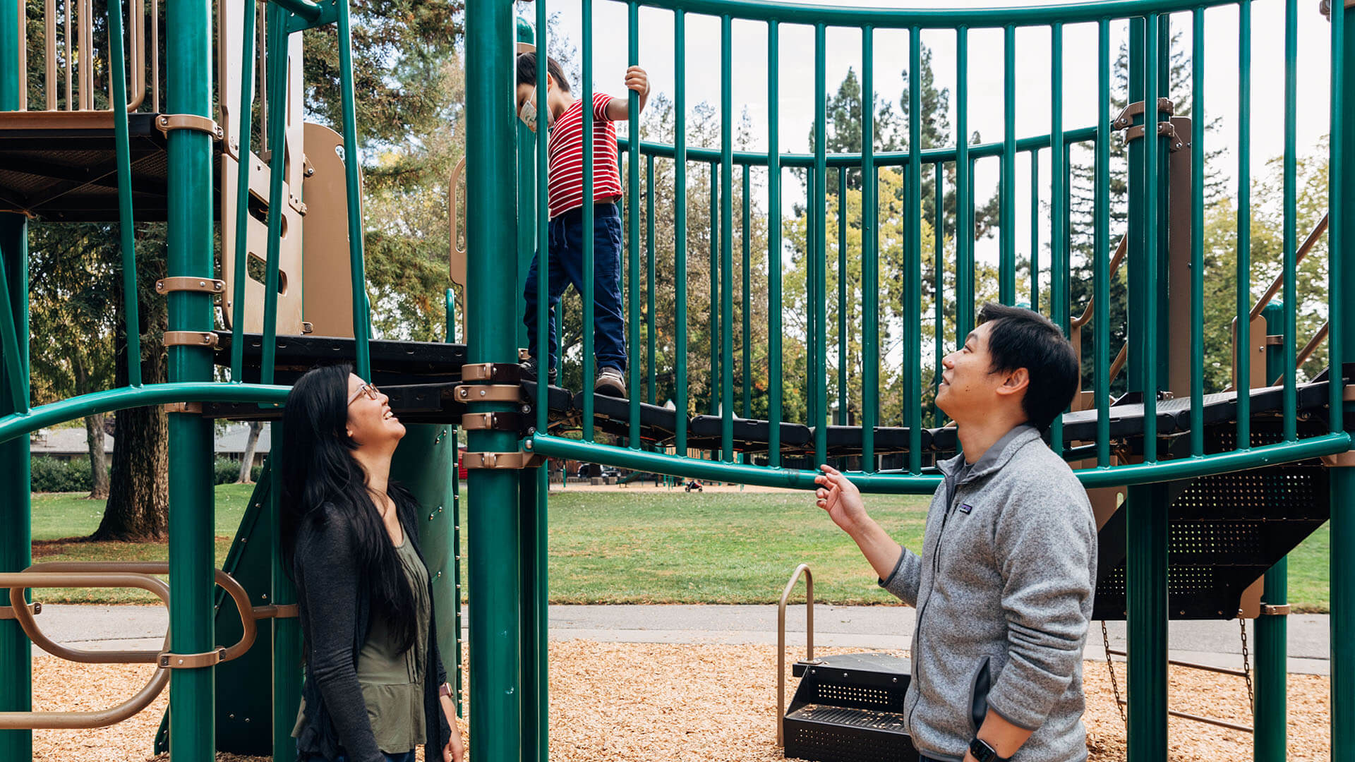 A man and women looks up at their son who is walking across the bridge of a playground.