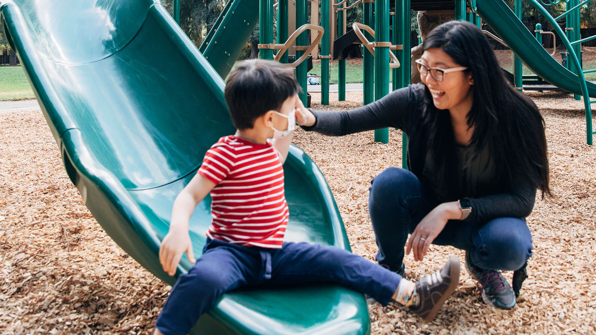 A boy wearing a mask sits at the bottom of a playground slide looking at his mother who is kneeling next to him with a smile.