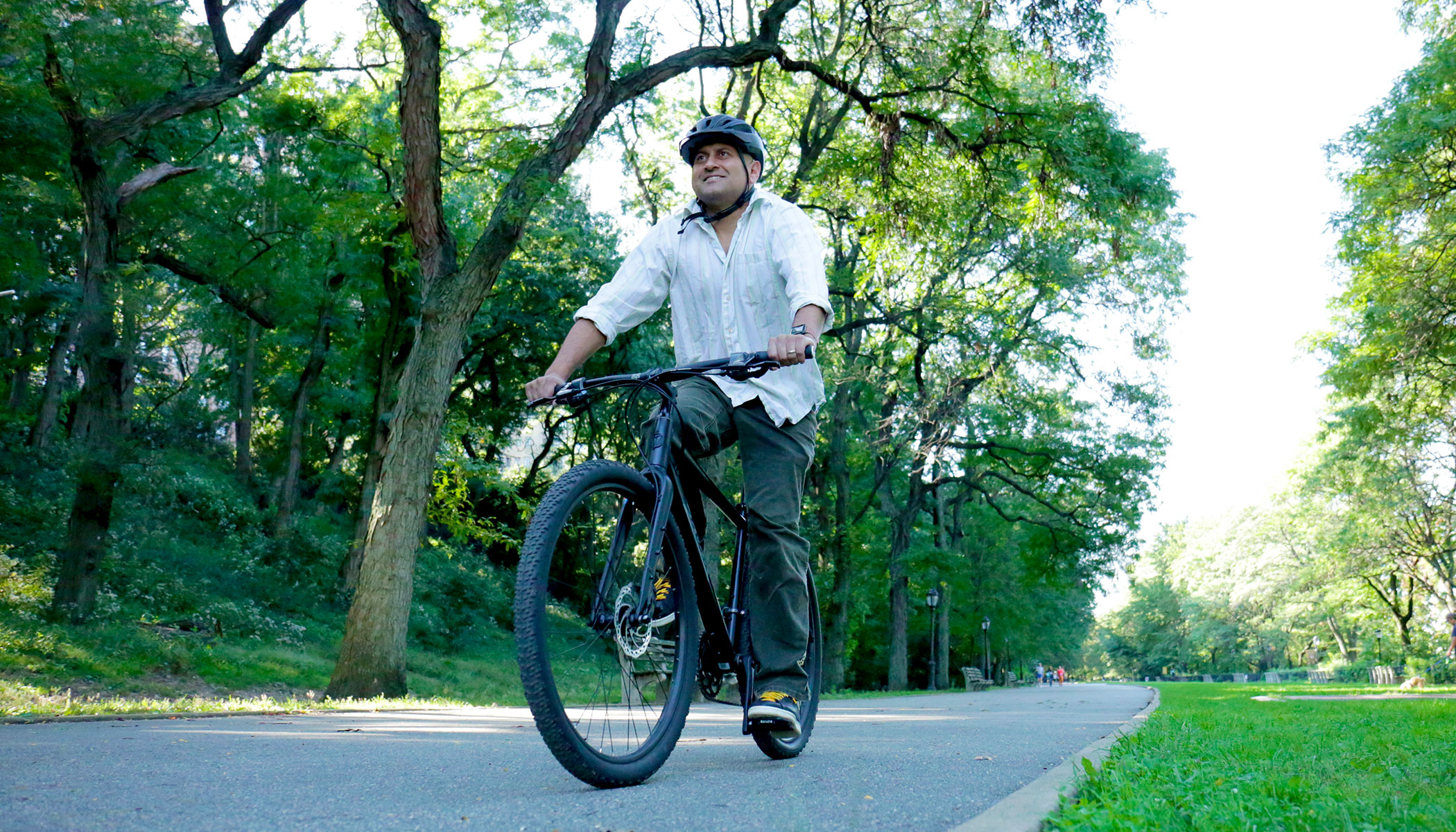A man rides a bike wearing a helmet. Green trees are in the background.