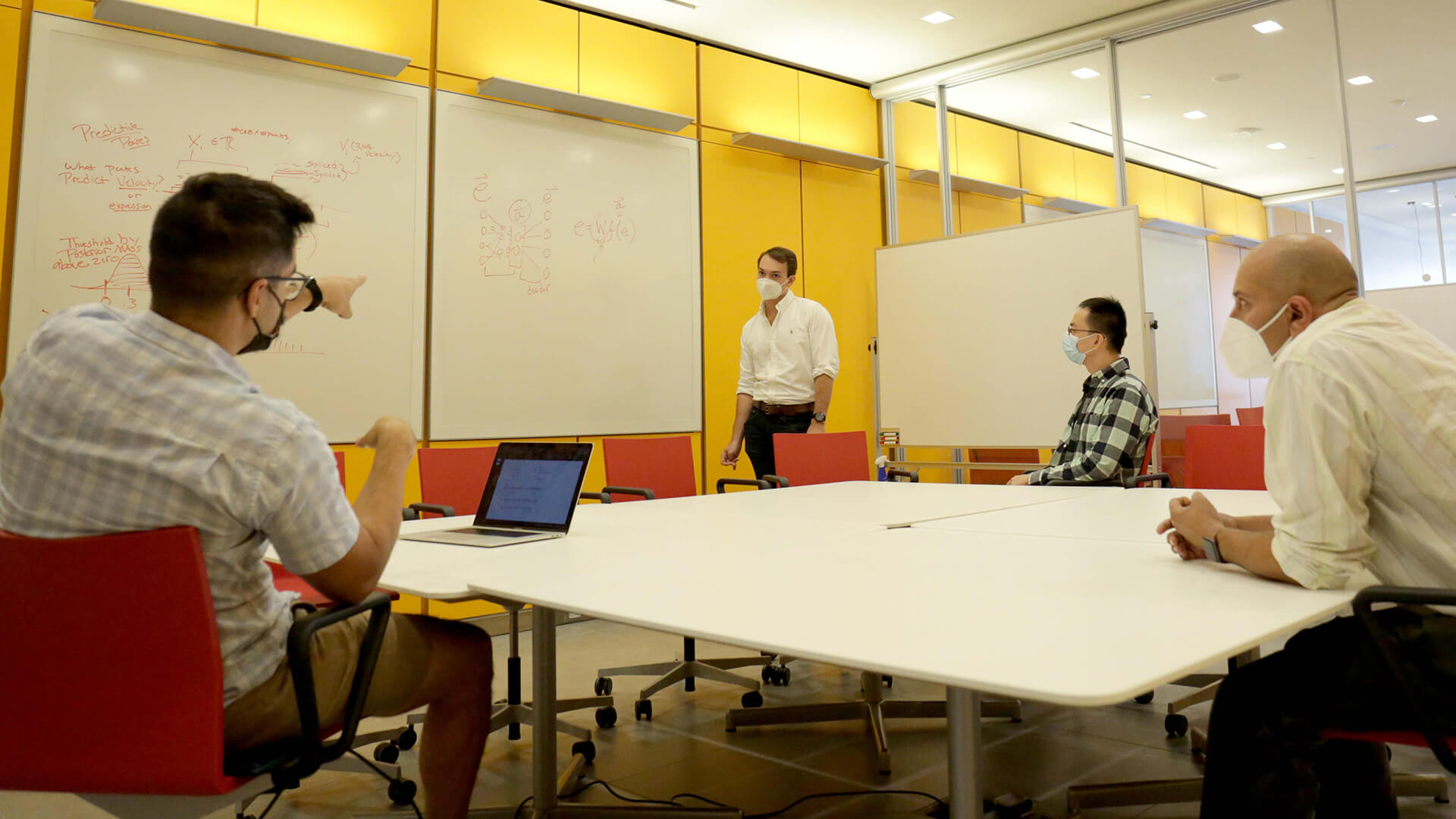 A group of people wearing masks discuss the content of a whiteboard in a meeting room.