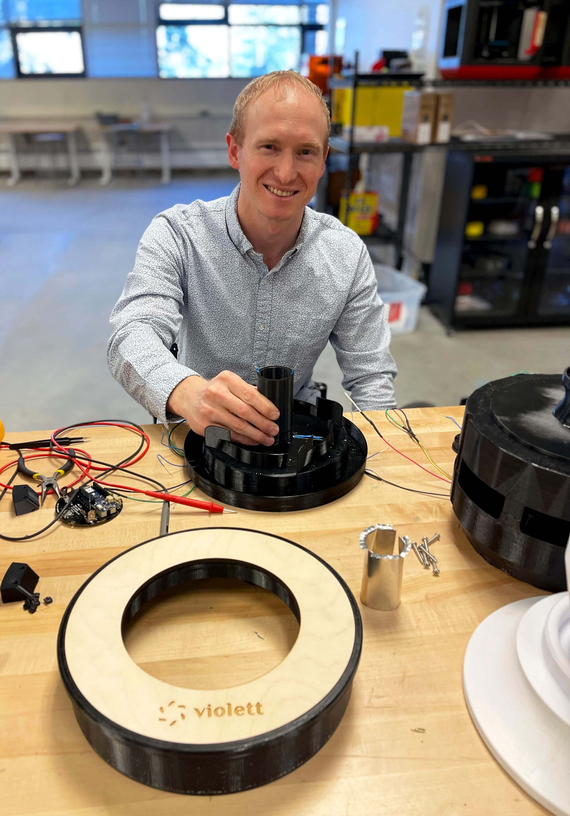 A man smiles as he sits at a work table with electrical components. 