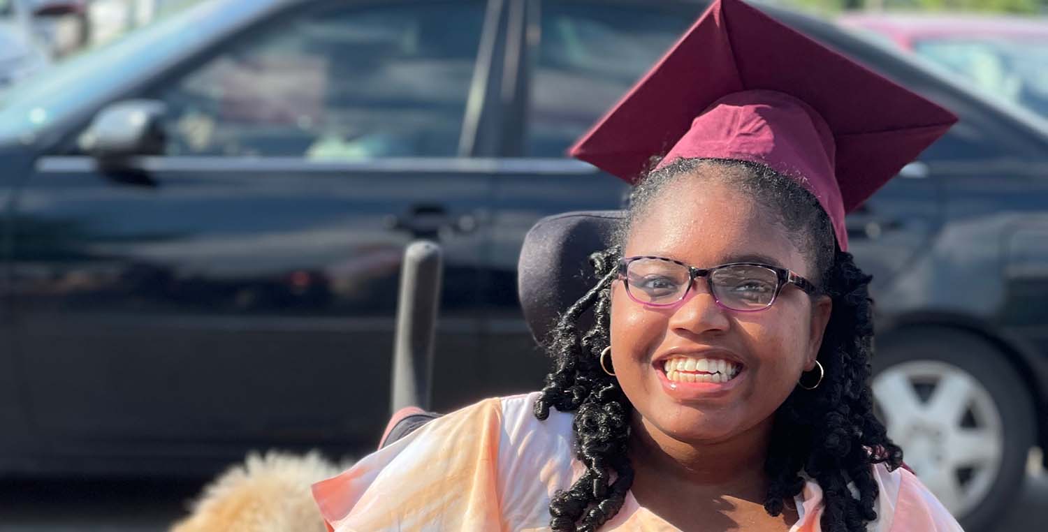 Khari McCrary, a teenage girl, wearing a graduation cap and sitting in a wheelchair smiles with her seizure service dog in the school parking lot.