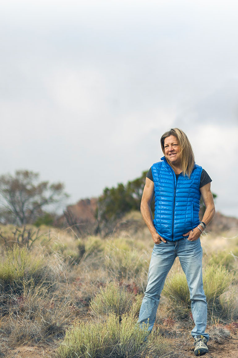A woman wearing blue jeans and a puffy vest smiles and stands in a field with her hands in her pockets.