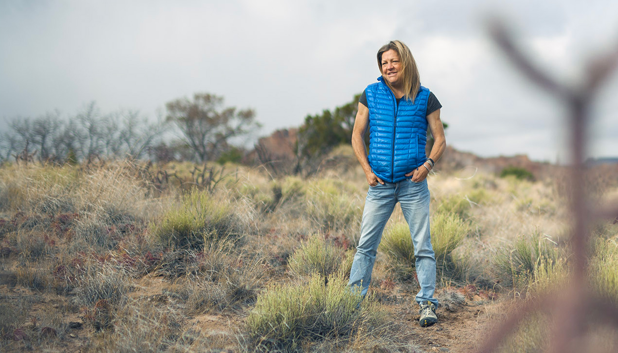 A woman wearing blue jeans and a puffy vest smiles and stands in a field with her hands in her pockets.