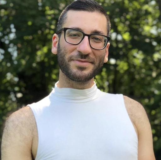 A queer, genderqueer, arab artist and activist with glasses, a trim beard and a crew cut in a white mock turtle neck. They are standing confidently in front of trees in a park.