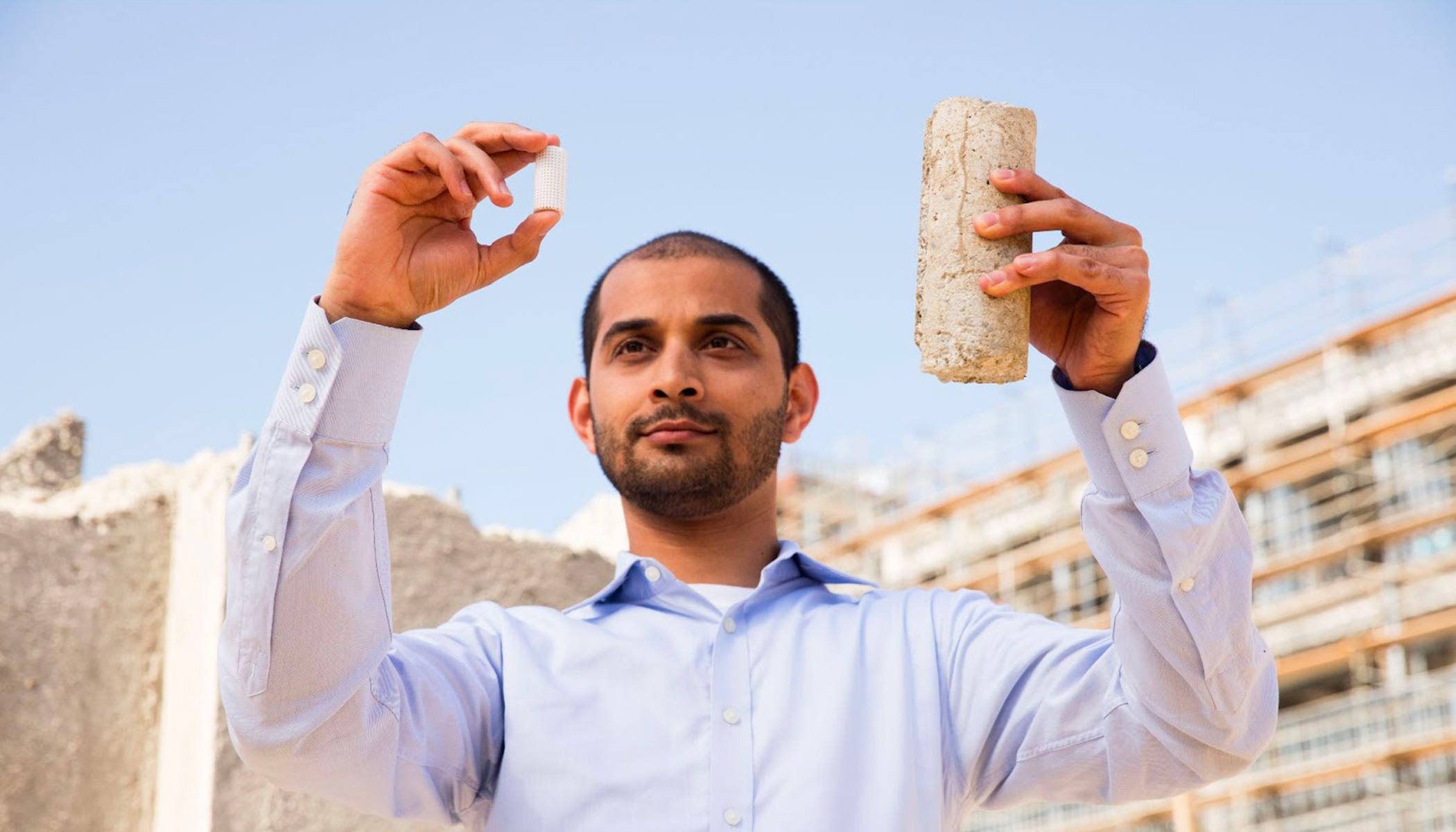 Gaurav, in blue shirt, holds a thimble-sized piece of low-carbon concrete in his left hand and a bottle-sized piece of concrete in his right hand.