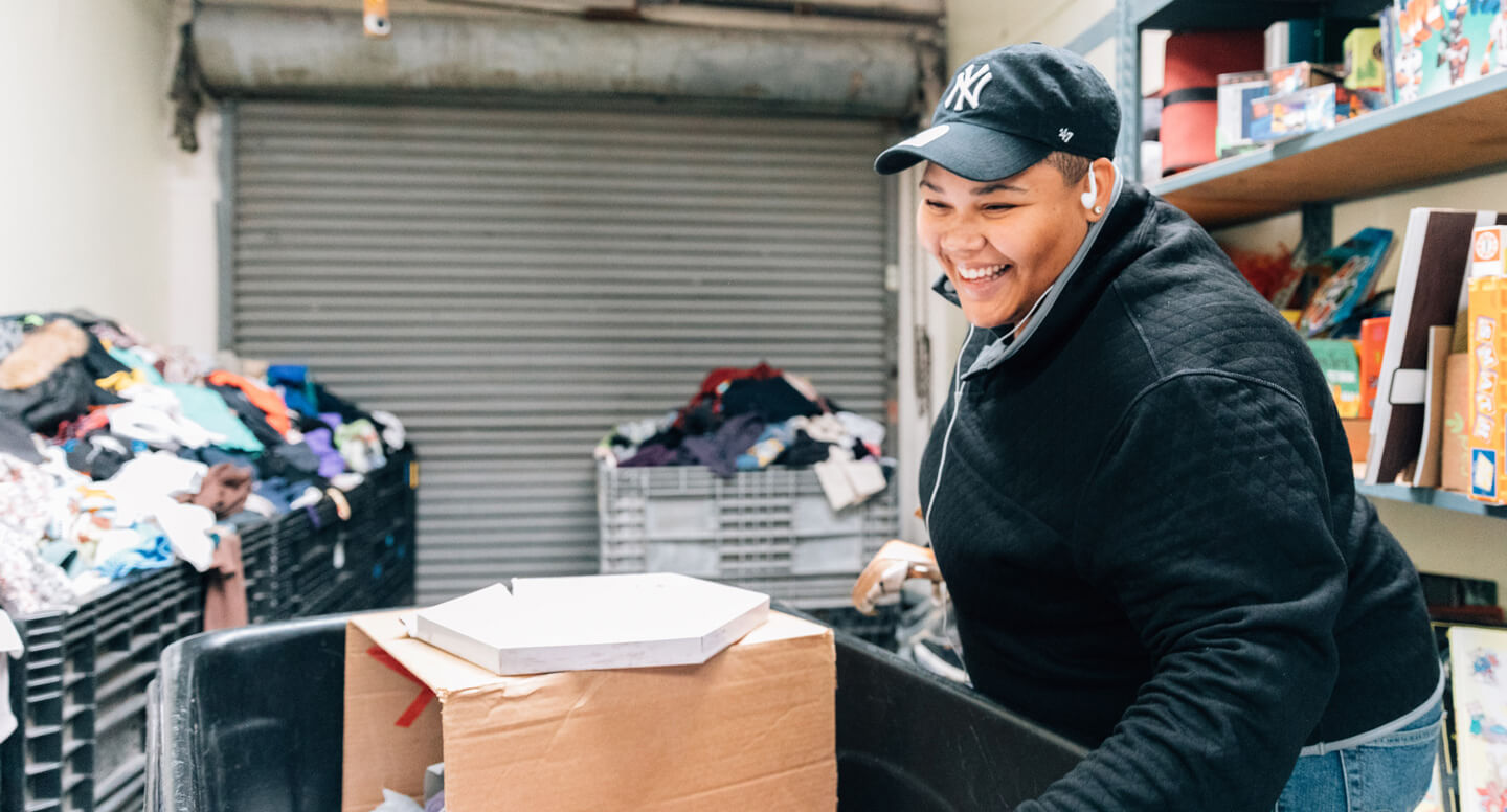 Smiling woman in black jacket and New York Yankees hat sorting donated goods and clothing at Ecumenical Hunger Program.