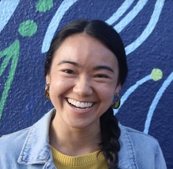 East Asian woman wearing earrings and smiling. She is wearing a yellow blouse and blue jean jacket.