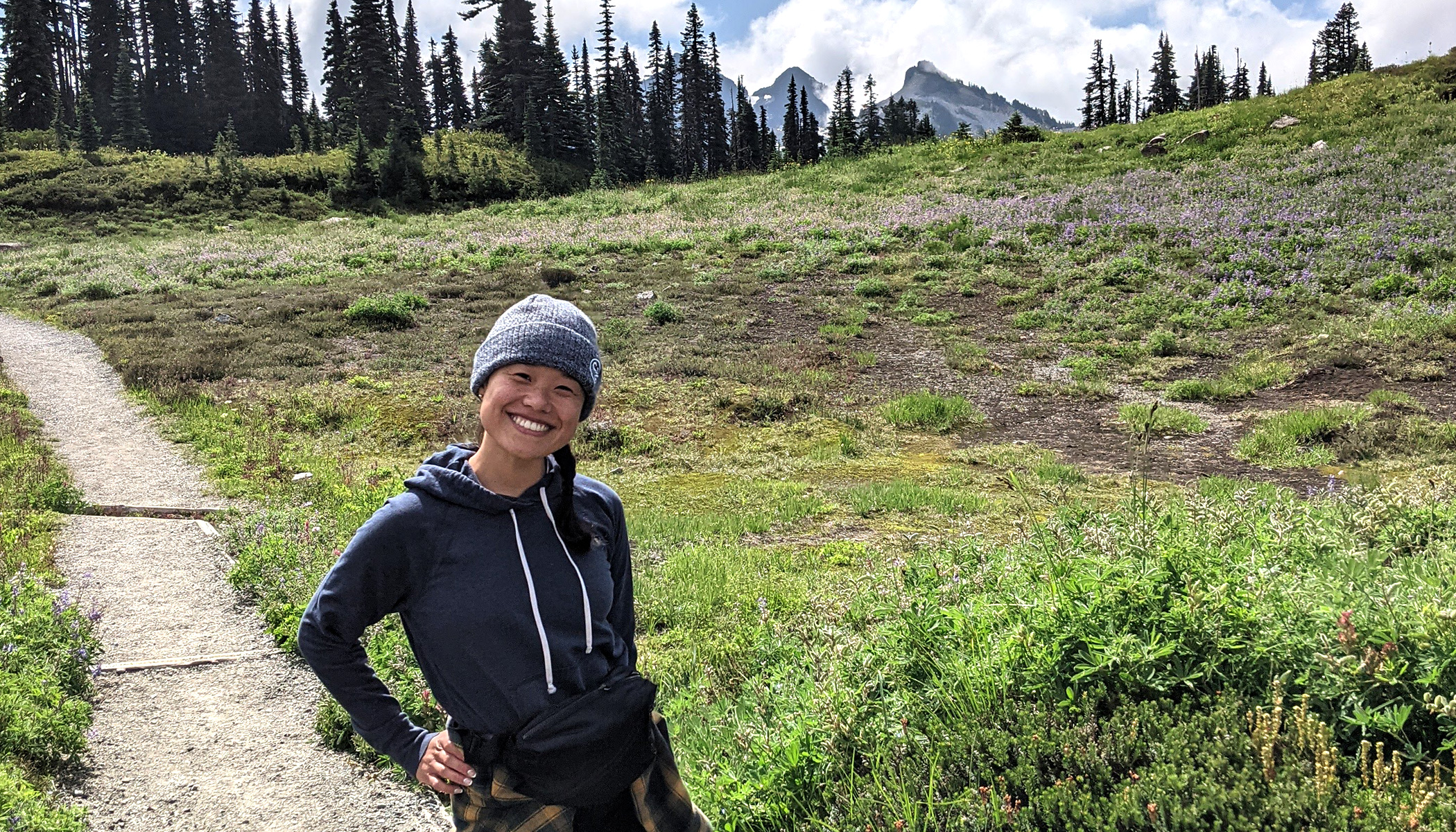 CZI housing affordability manager Michelle smiles as she stands on a trail, surrounded by greenery. You can see the mountains deep in the background.