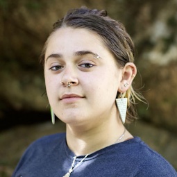 An image of Stefanie, a white non-binary person, slightly smiling in front of large brown stones, trees and green plants. They are wearing a blue t-shirt, long triangular earrings, and have several facial piercings.