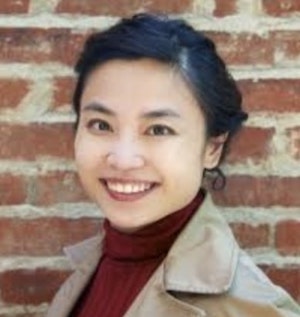 Asian-American woman with black hair and brown eyes wearing a red shirt and cream-colored jacket smiling in front of a brick wall.