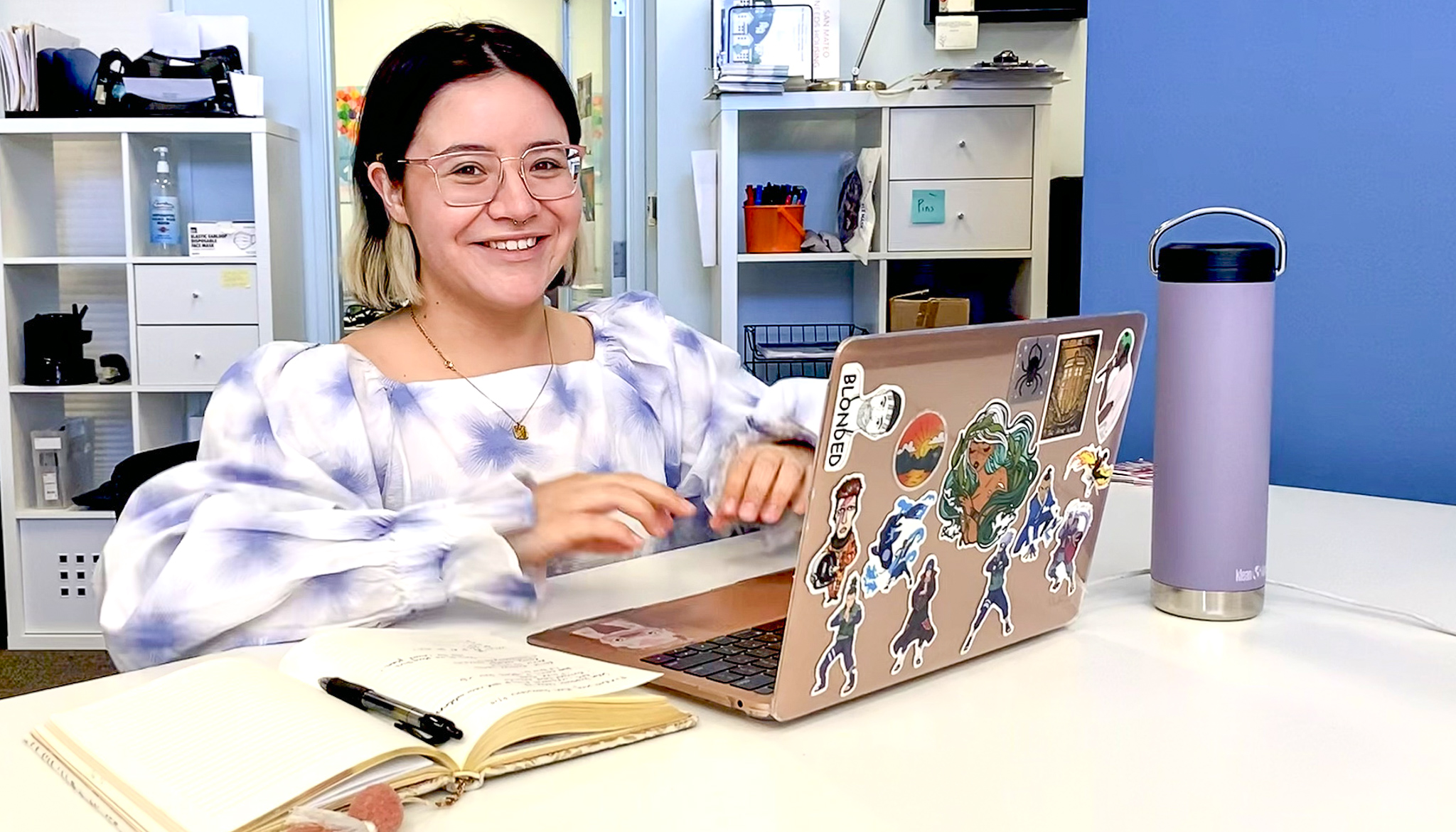 A woman with glasses wears a poofy, purple and white blouse and sits at a table with a laptop covered in stickers.