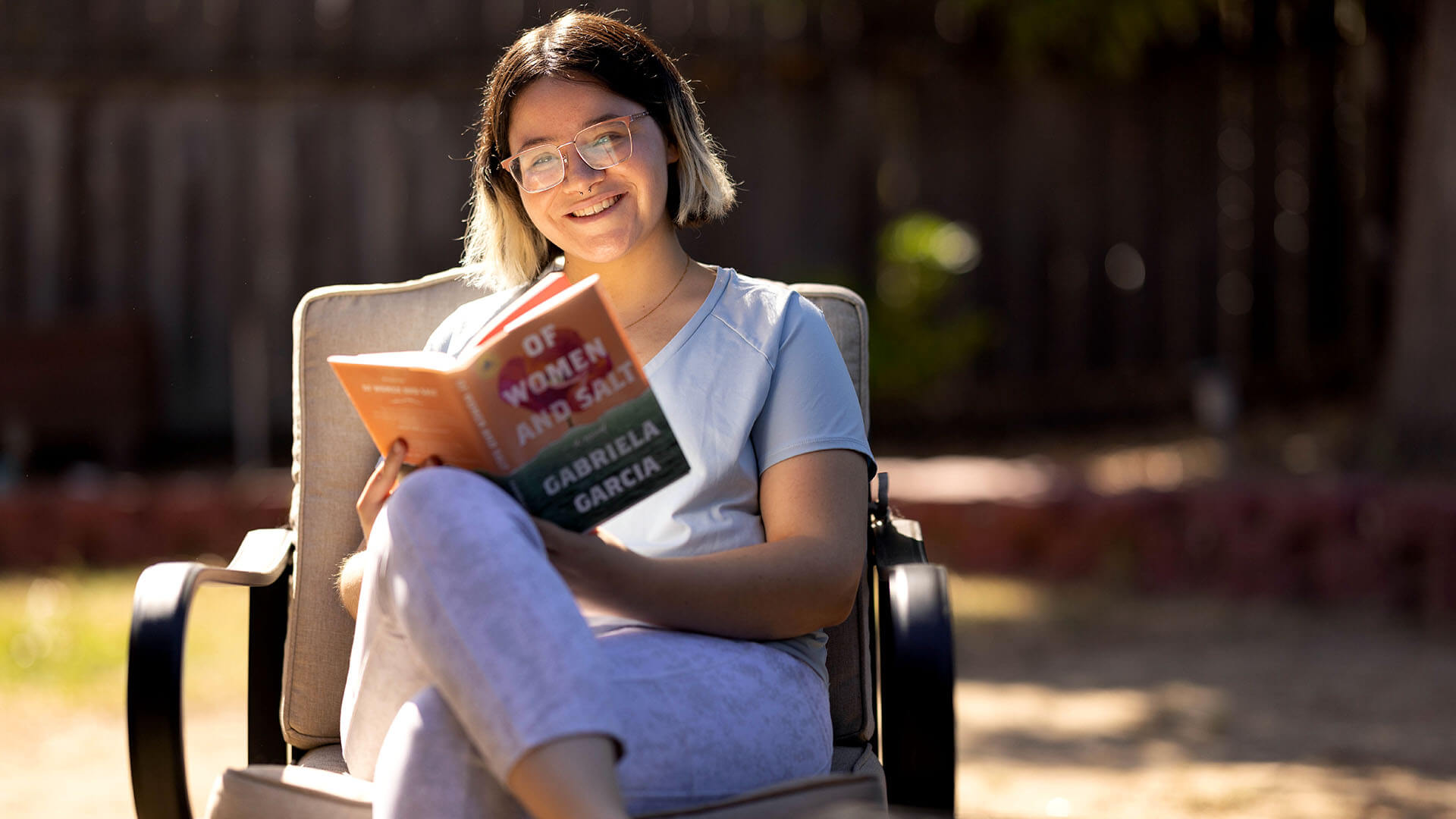 A woman with glasses smiles at the camera while holding a book titled “Of Women and Salt.” 