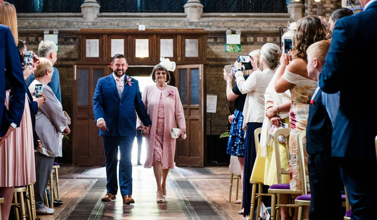 Glyn in a blue suit smiling and walking down the aisle while holding hands with his mother.
