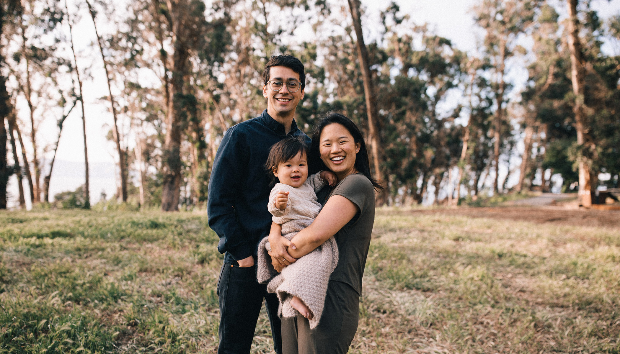 Julio, his spouse and child smile for a picture, surrounded by grass and trees.