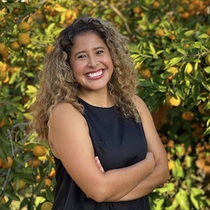 Anna, a Latinx non-binary person with brown skin, long curly brown hair with blonde highlights, smiling and standing in front of a mandarin tree in full bloom