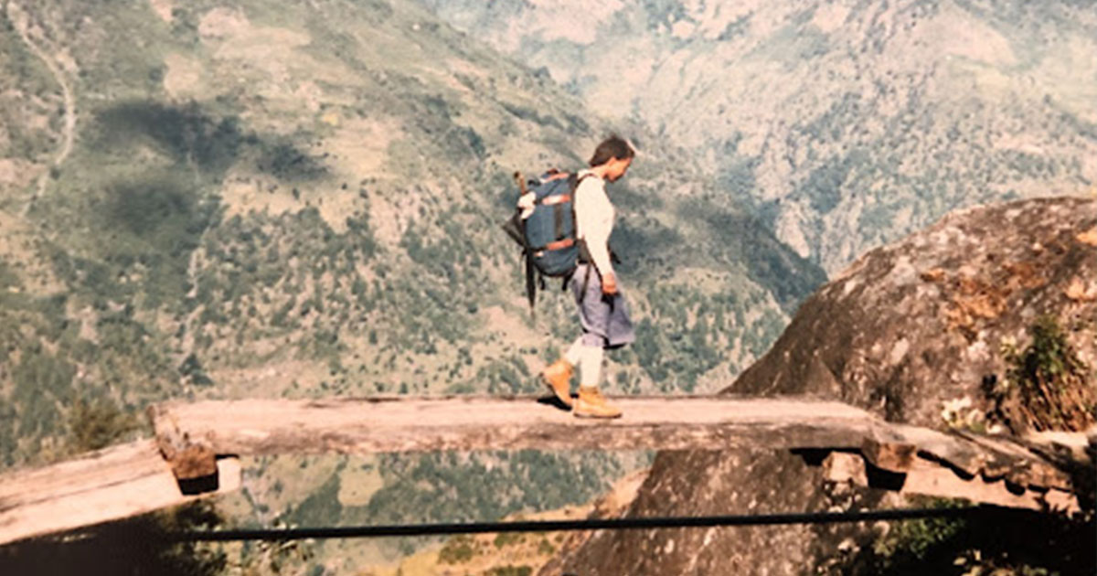 A woman wearing hiking boots and a backpack walks on a small bridge in Nepal. Mountains are in the background.