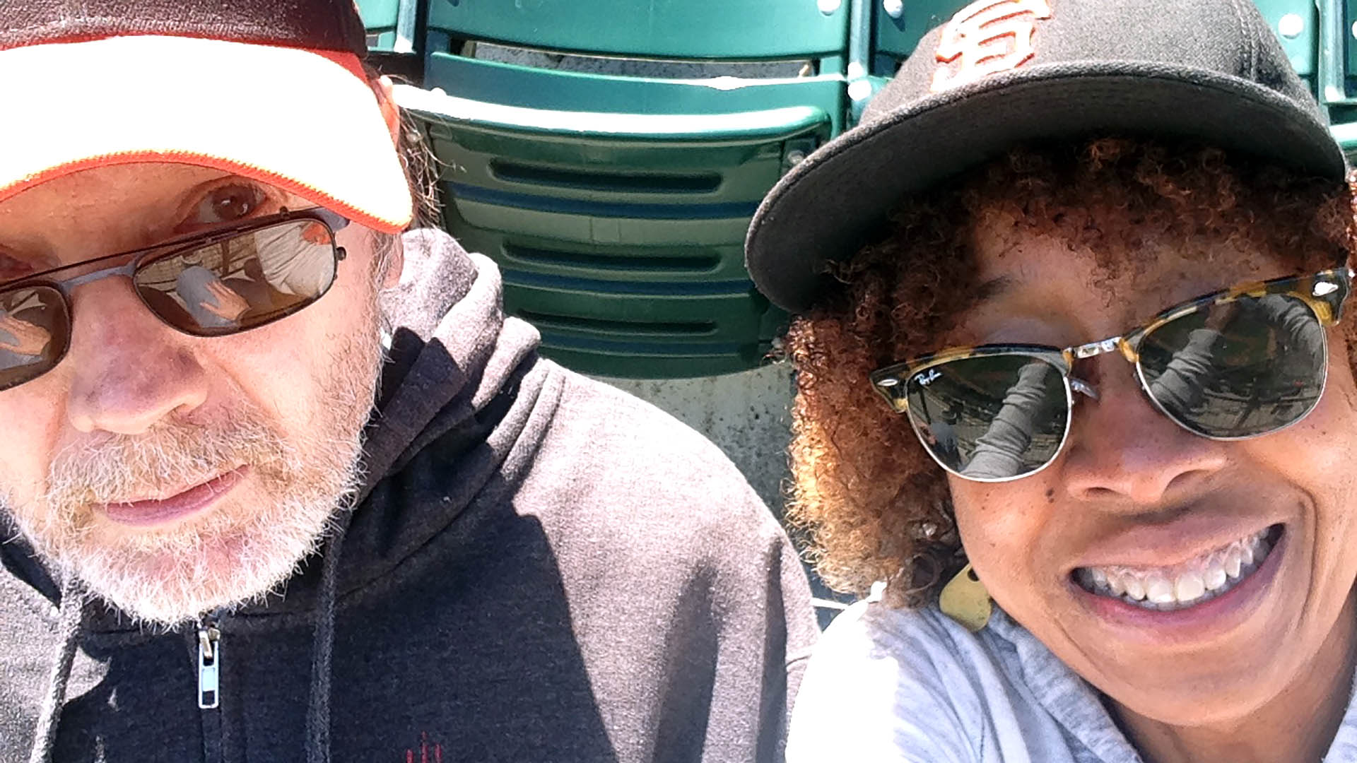 A man and woman wear baseball caps and sunglasses in a stadium.