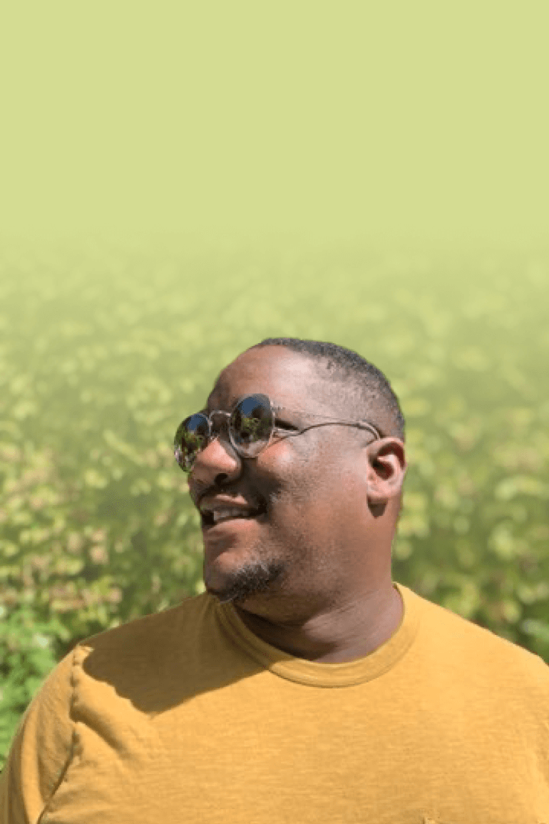 Dakarai wears a yellow shirt and sunglasses while smiling for an outdoor photo.