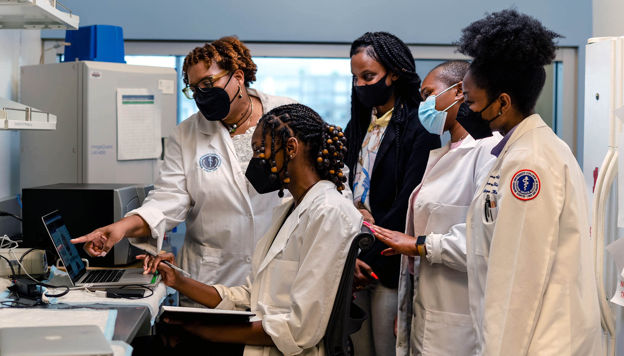 A group of Howard women researchers wearing lab coats look at a laptop in a lab.