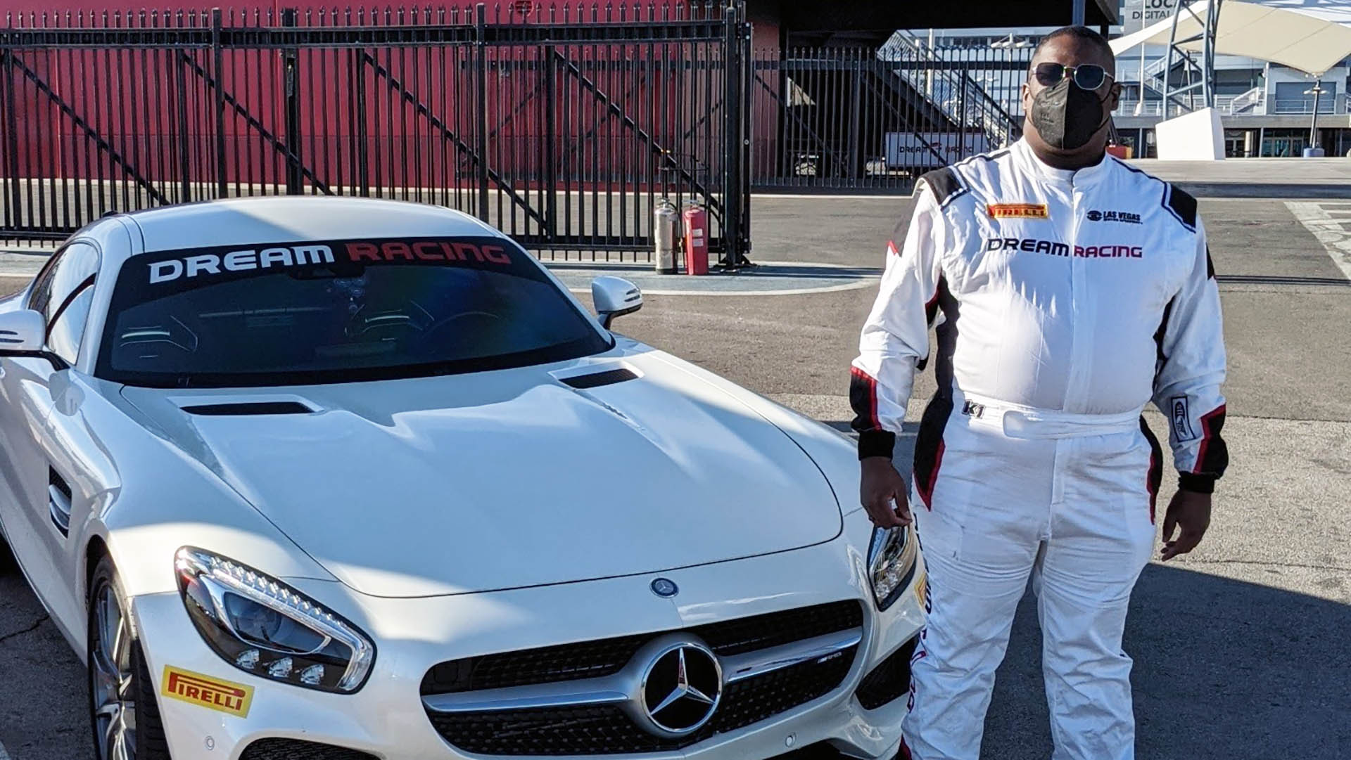 Dakarai wears a mask, sunglasses, and a racing uniform while standing next to a Mercedes that says “Dream Racing” on the windshield.