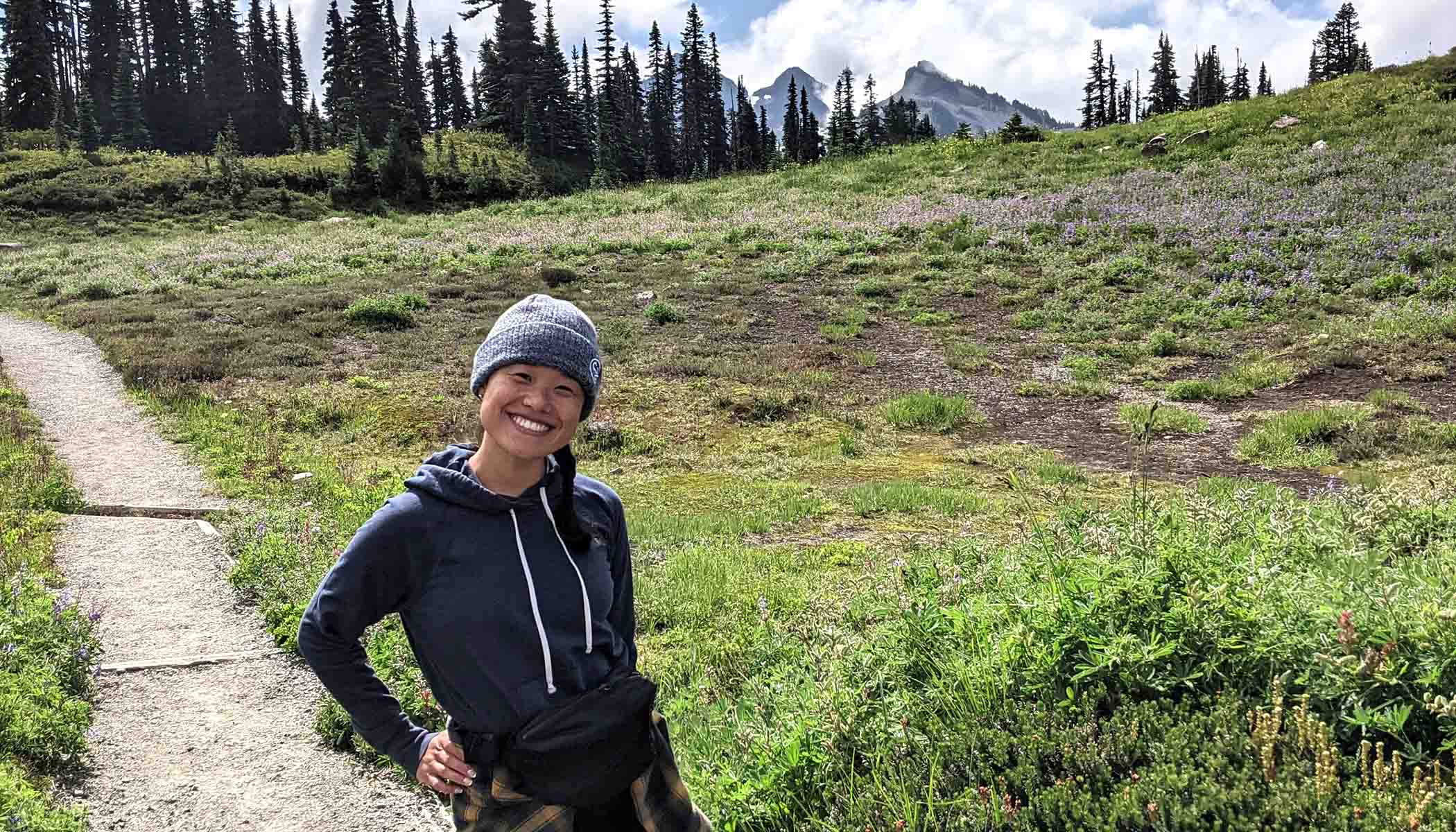 A woman smiles while on a hike. Greenery, trees and mountains are in the background.