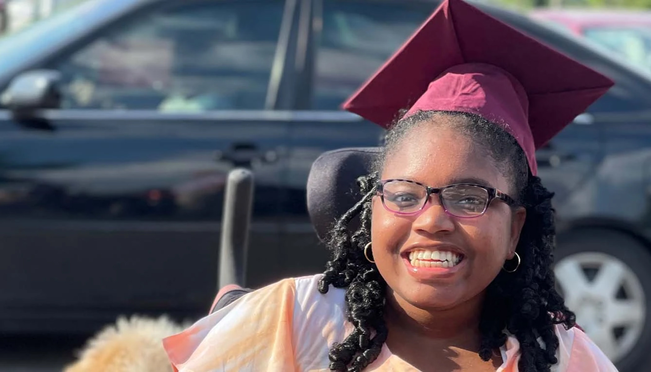 Khari McCrary, a teenage girl, wearing a graduation cap and sitting in a wheelchair, smiles in the school parking lot.