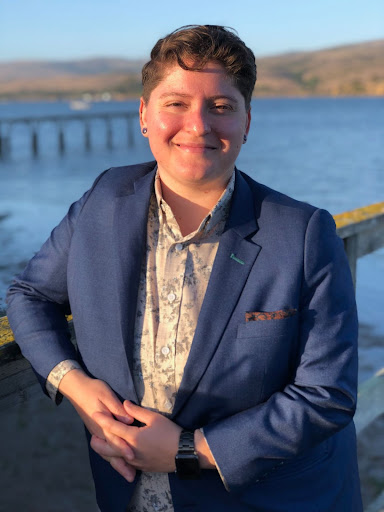 A white, queer person with short brown hair and a dapper navy suit. They are smiling on a dock next to a bay.