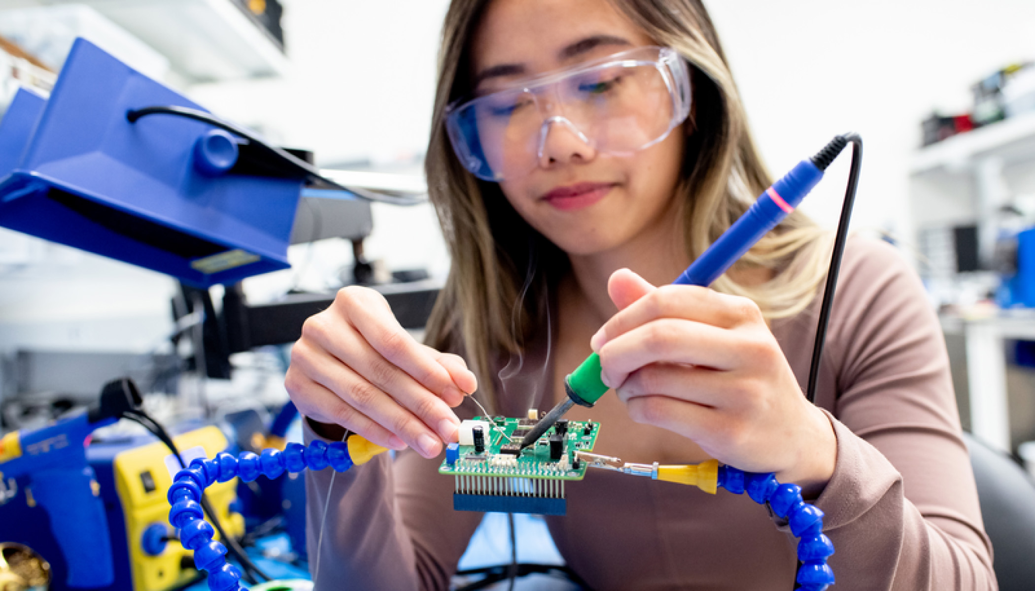 A scientist in a lab wearing clear safety glasses uses both hands to work on a circuit board.