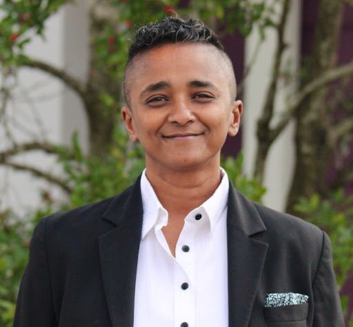 A masculine of center woman of color with short curly hair is smiling at the camera. She is wearing a white button down shirt with black buttons, a black blazer and a blue and white floral pocket square. There is blurred greenery in the background.