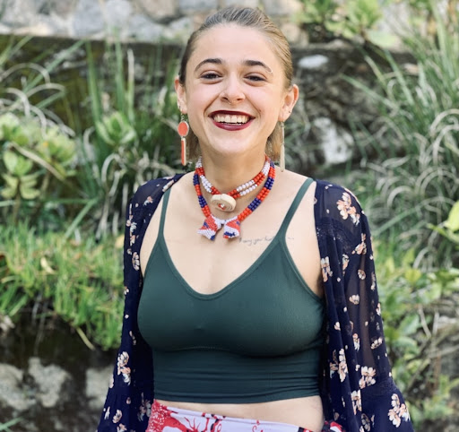Stefanie, a white, non-binary person, stands smiling in front of an array of green plants and stones. They are wearing a blue floral cardigan, a dark green top, and red, blue, and white beaded necklaces. They have several facial piercings, long, dangly red earrings, and are wearing maroon lipstick.