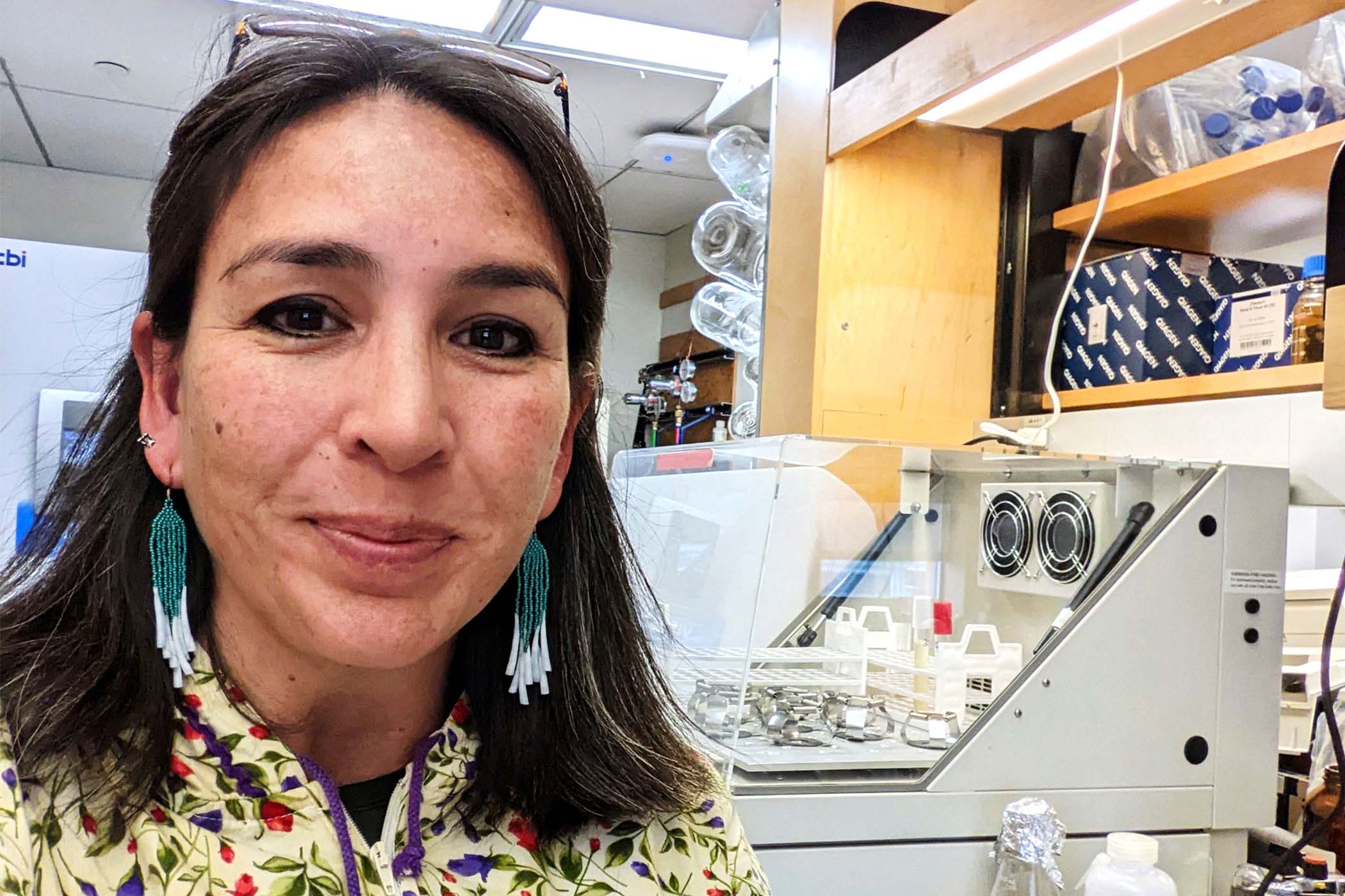 Dr. Kat Milligan-McClellan smiles directly at the camera. She is wearing a floral top. In the background is lab equipment, including test tubes.