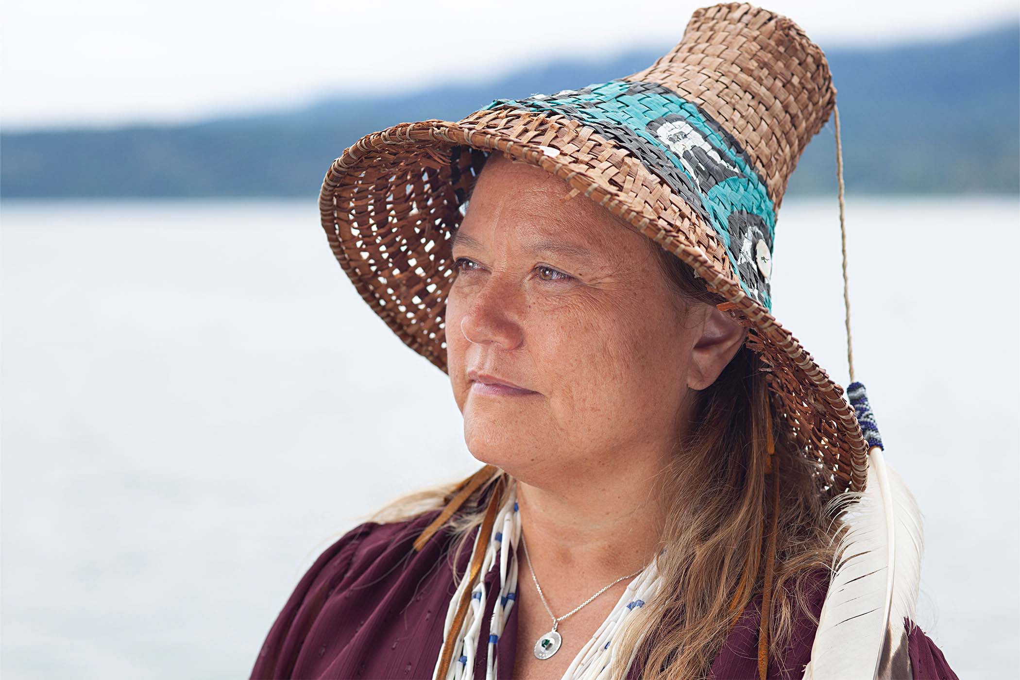 Fawn Sharp wearing a straw hat and maroon top gazes towards the left with serene expression. In the background, out of focus, is a view of a lake and mountains.