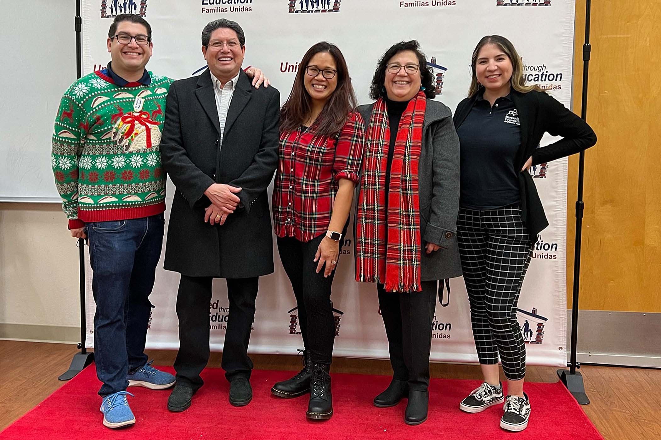 Two men (left) and three women (right) smile directly at camera. Most are in holiday attire and all are standing at a step and repeat.
