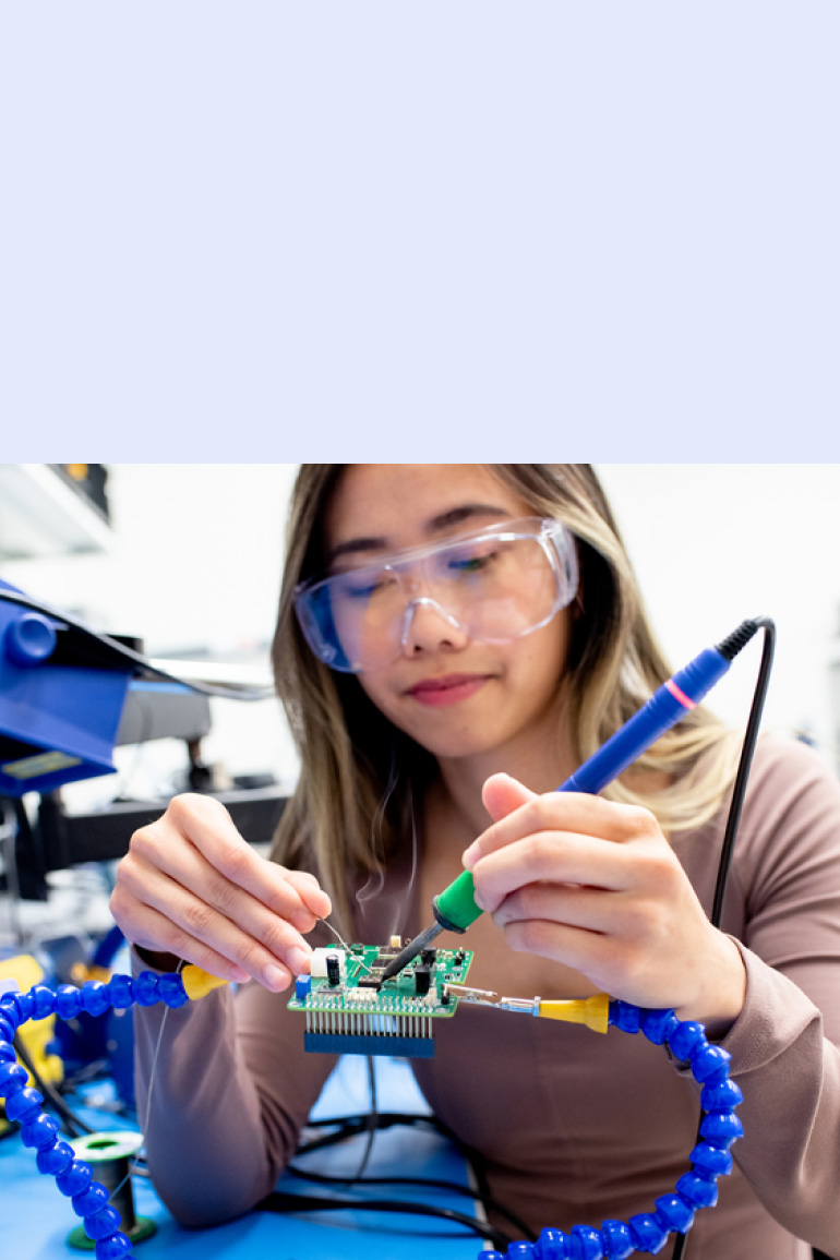 A scientist in a lab wearing clear safety glasses uses both hands to work on a circuit board.