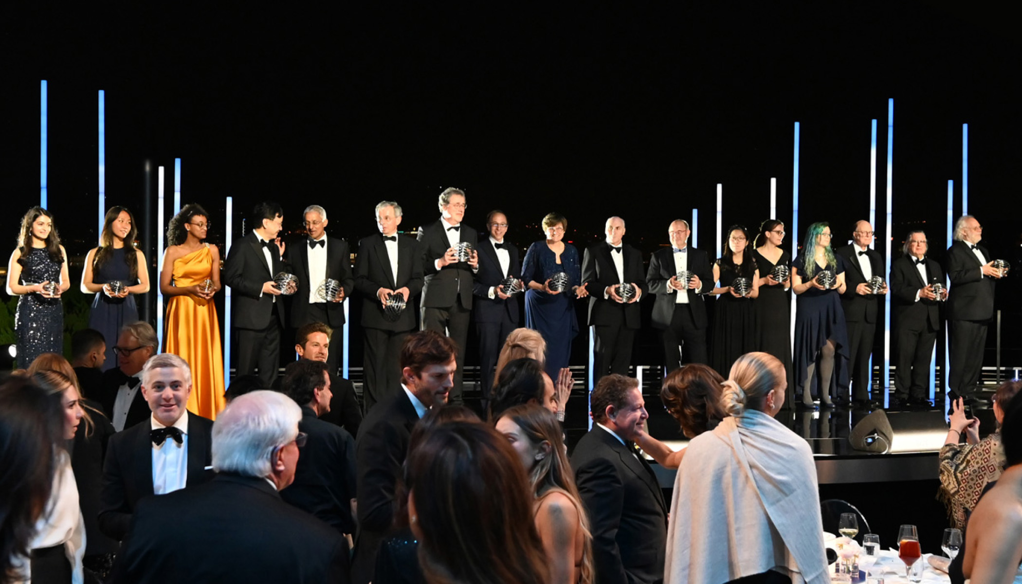 A large group of people stand onstage holding a spherical award in front of a crowd at the 9th annual Breakthrough Prize ceremony