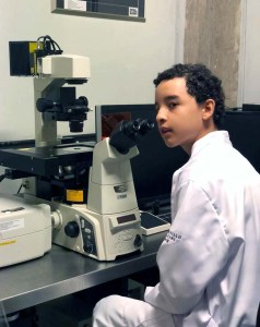 A young boy wears a white lab coat while using a large microscope in a lab.