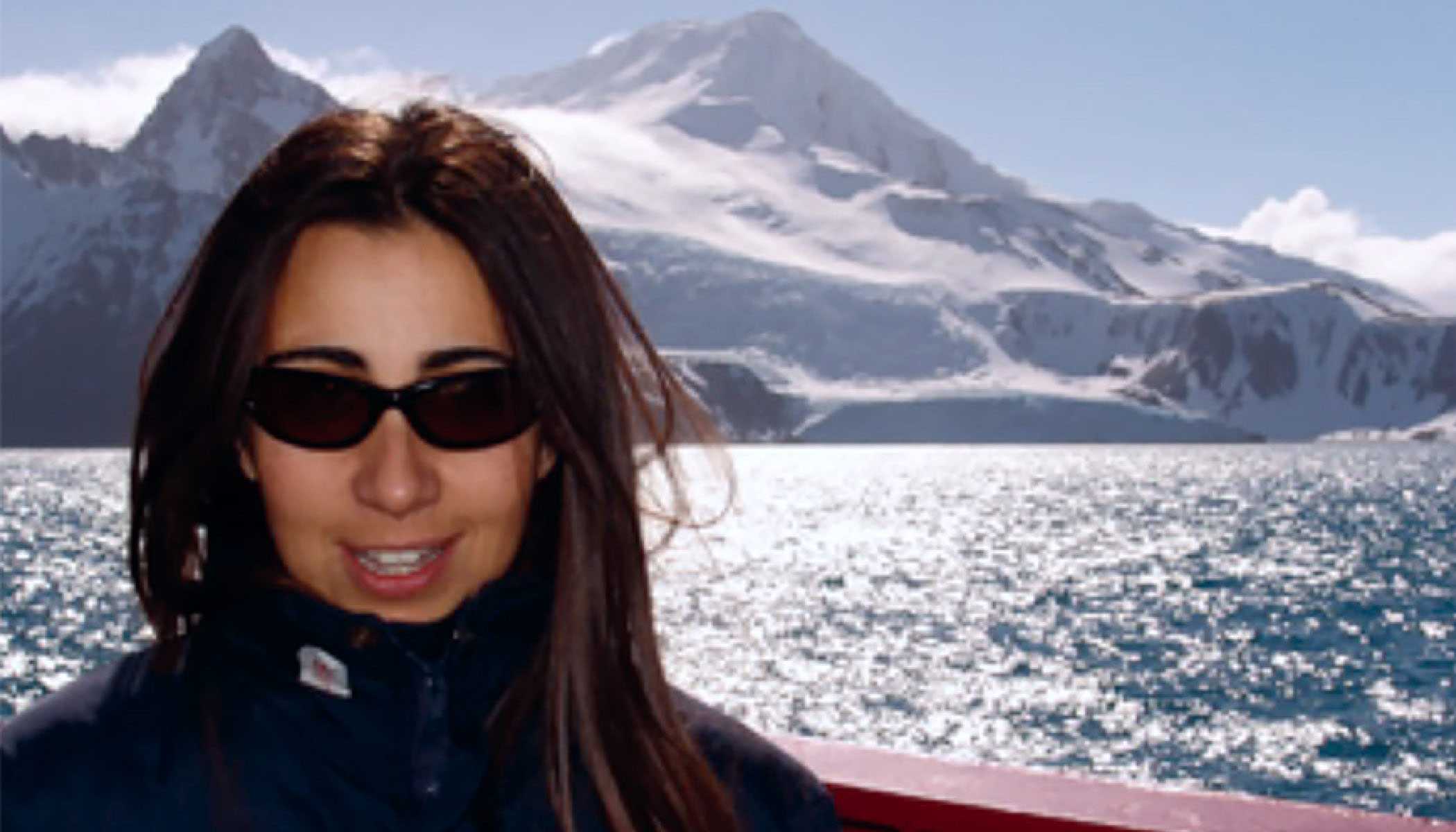 Scientist Débora Iglesias-Rodriguez wearing sunglasses and black coat smiles. In the background is a mountainscape and body of water.