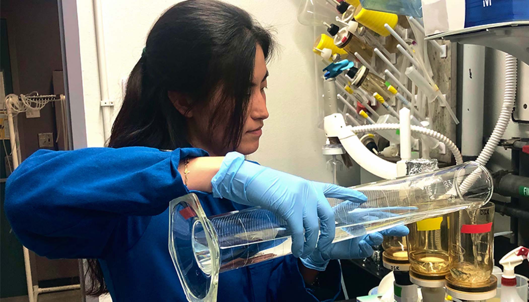Lab specialist Sylvia Kim wearing a blue lab coat and gloves pours a liquid from a glass beaker.