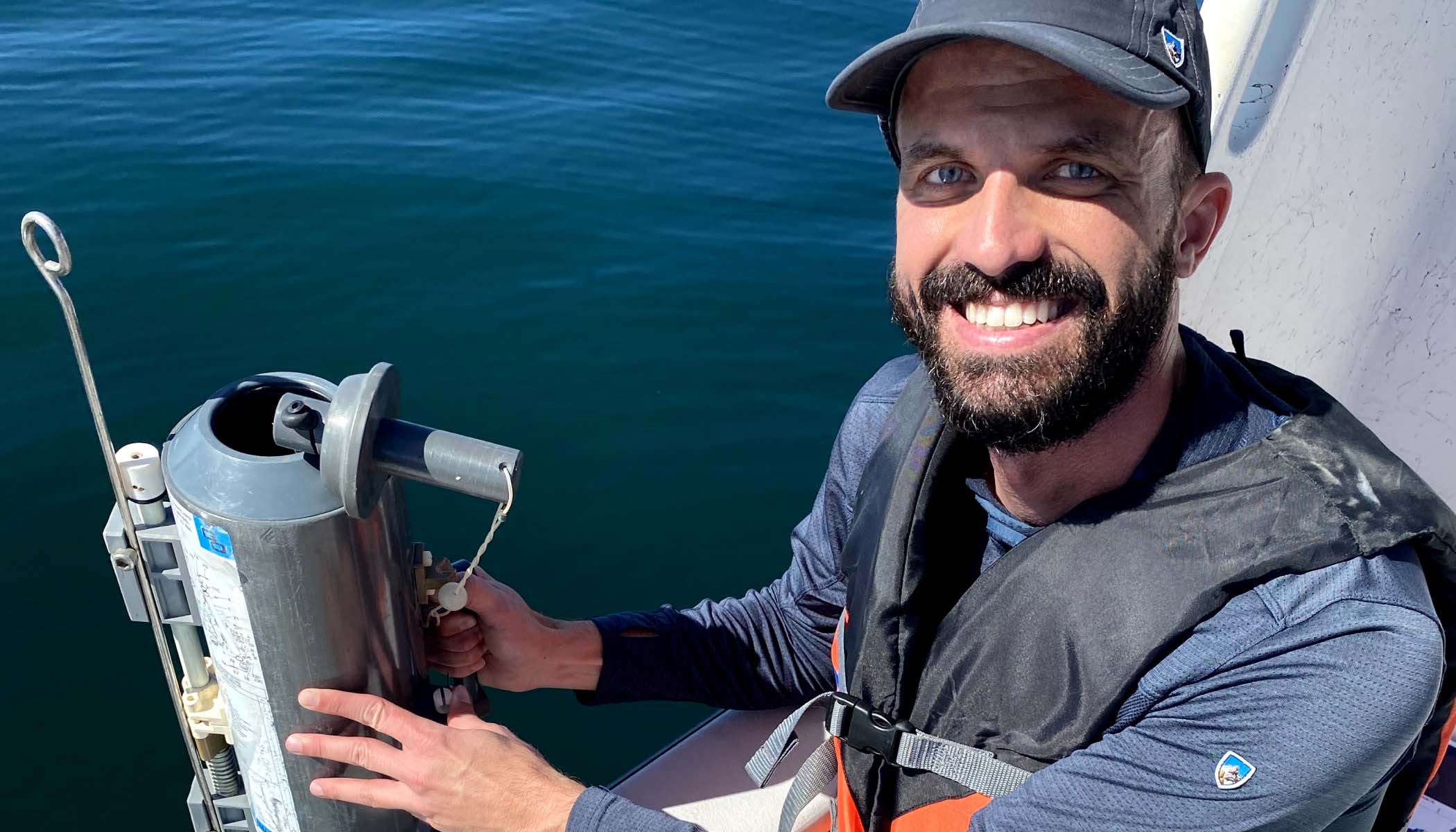 James Gately wearing hat and life vest sits in a boat, smiles as he collects seawater samples.
