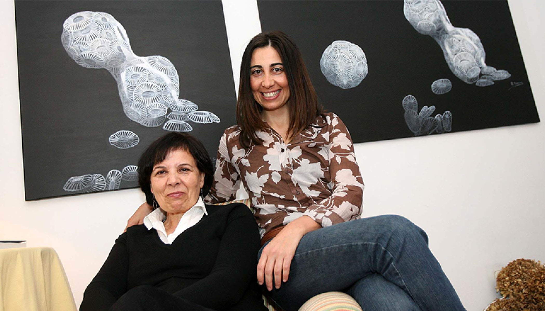 Two women sit side-by-side, the woman on the left wears a black sweater and white shirt, the woman on the right wears a brown, patterned shirt. On the wall behind them are two black and white paintings of coccolithophores.